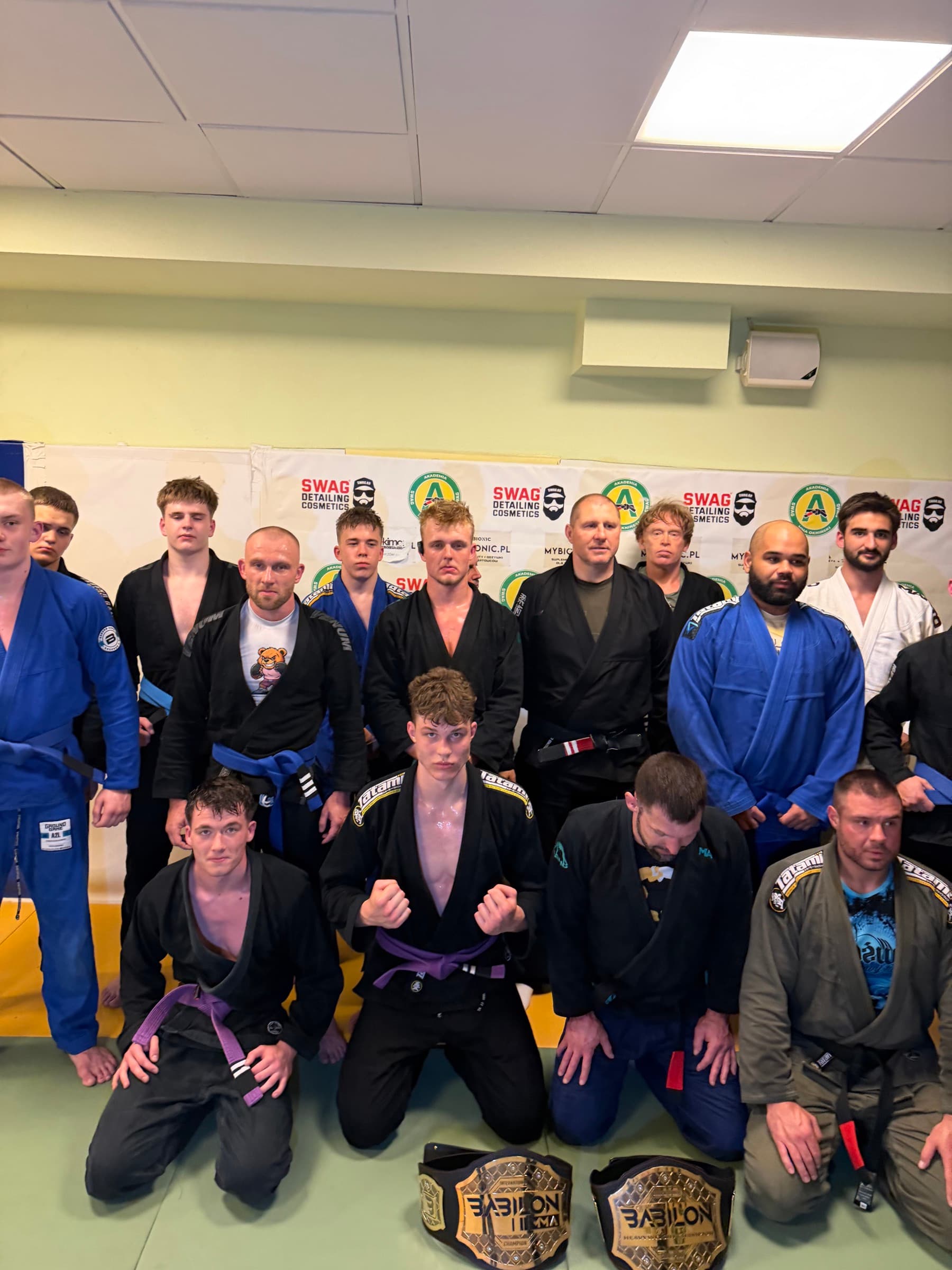 Grupa BJJ Akademii na macie, przed banerem klubowym, z pasami Babilon MMA