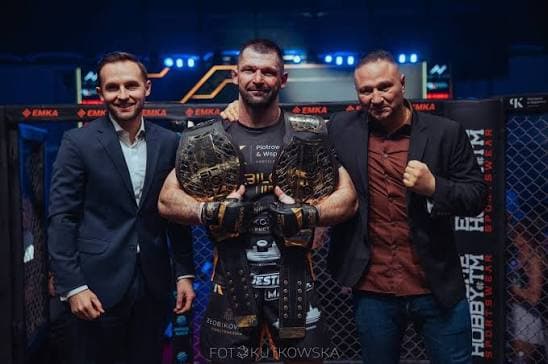 Szymon Kołecki w klatce z dwoma pasami mistrzowskimi Babilon MMA, obok dwie osoby ze sztabu