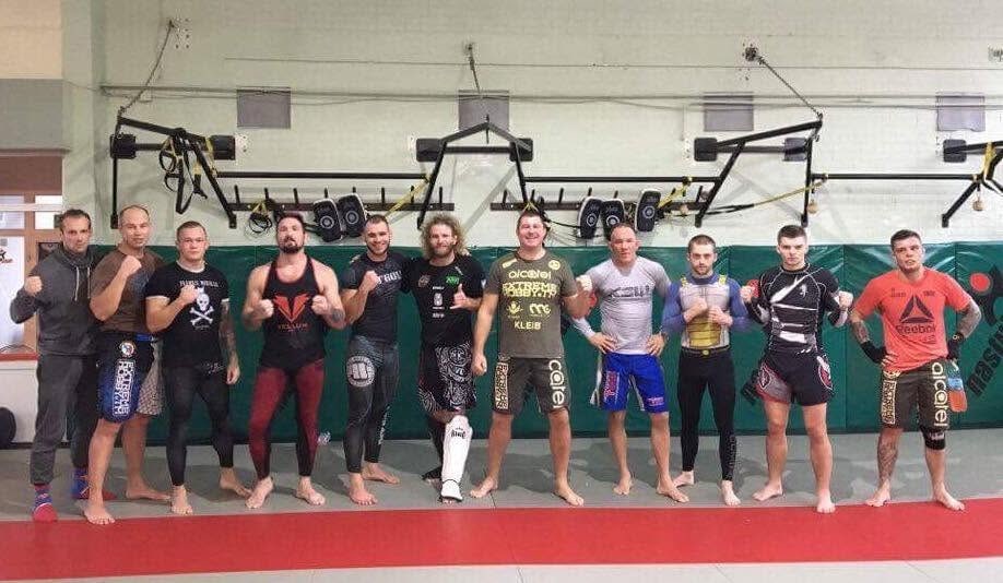 Grupa zawodnicza ustawiona w rzędzie w sali treningowej, w strojach do MMA