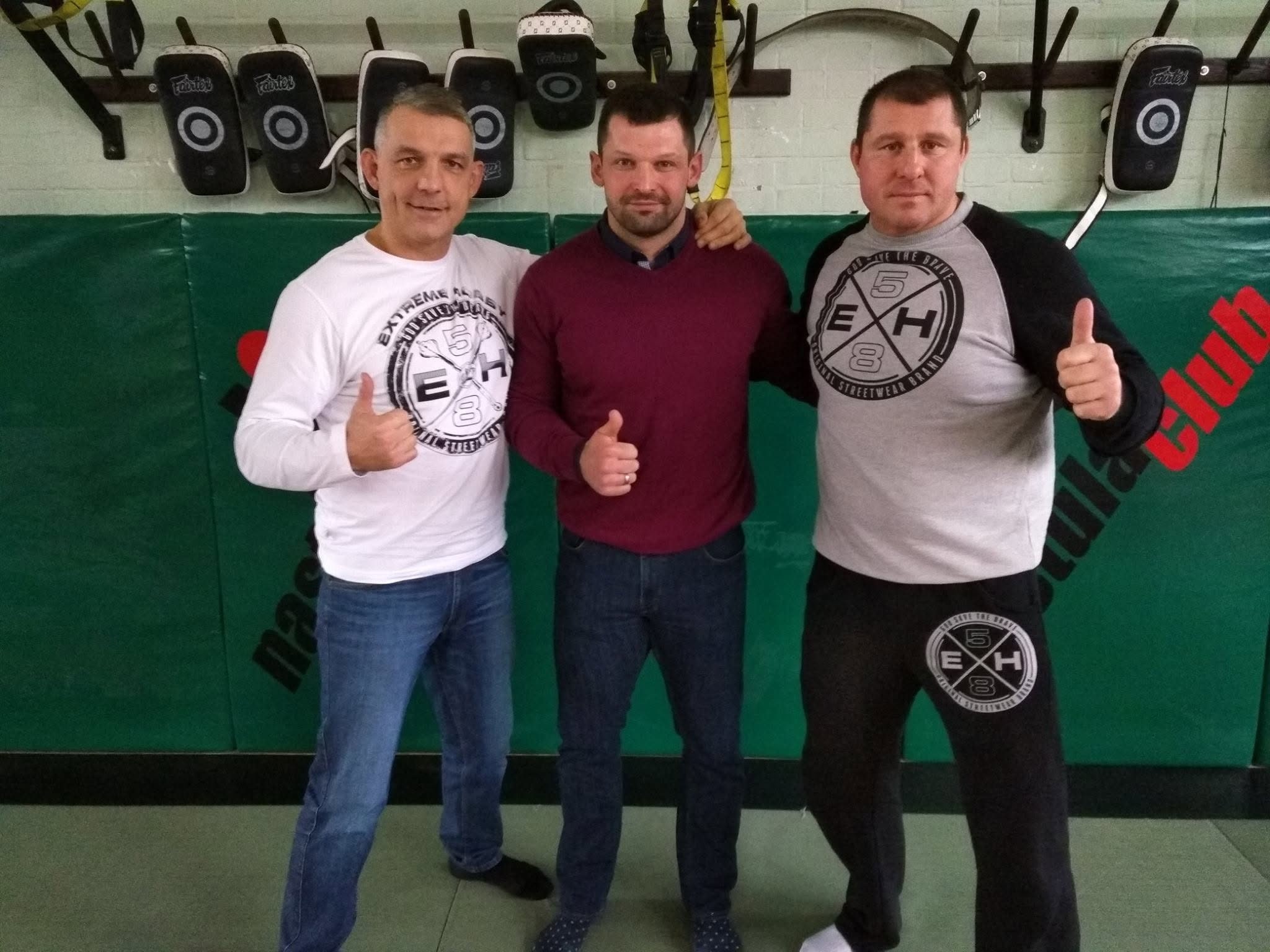 Trzej mężczyźni stoją obok siebie przy materacach w sali treningowej, dwaj pokazują kciuk w górę