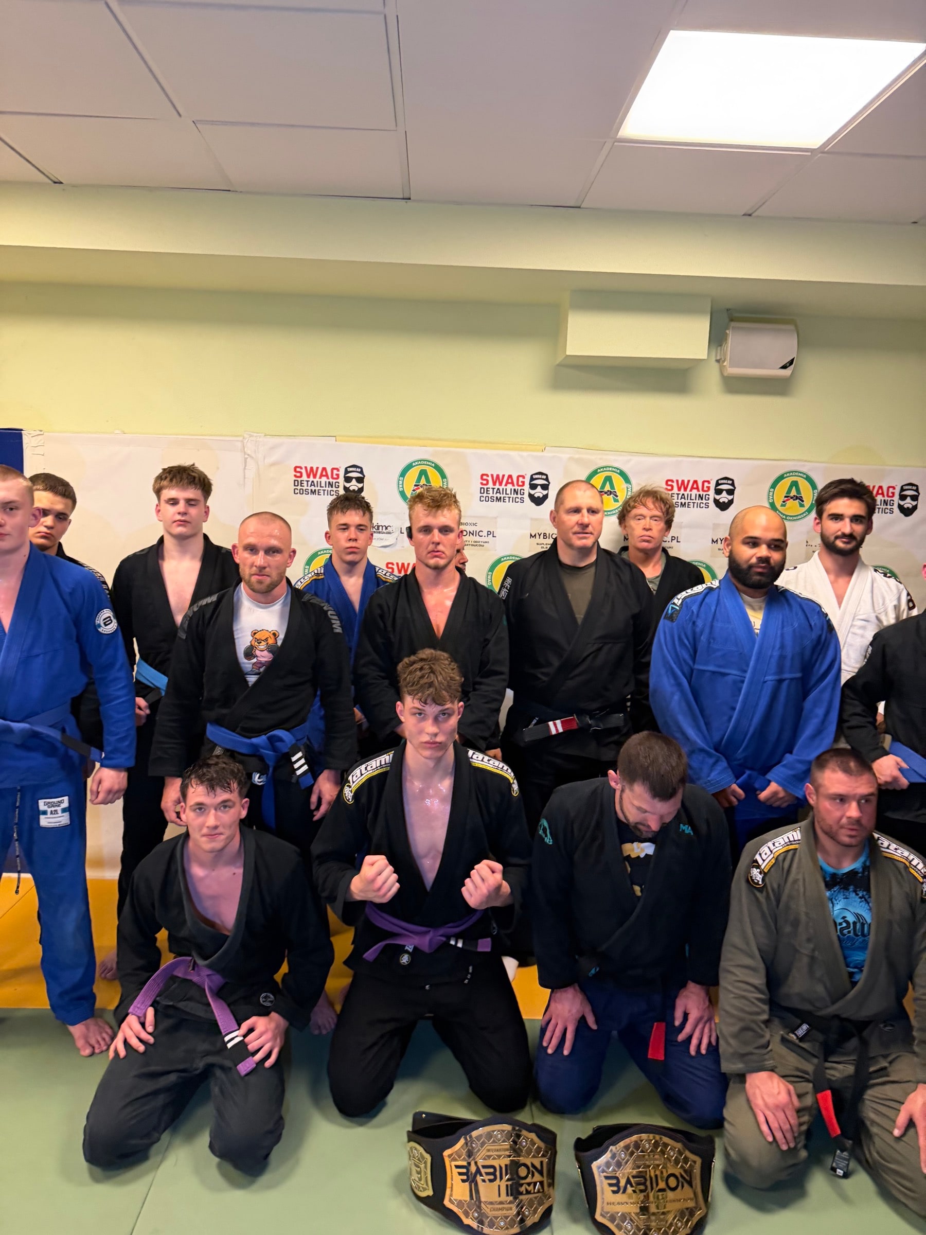 Grupa BJJ Akademii na macie, przed banerem klubowym, z pasami Babilon MMA