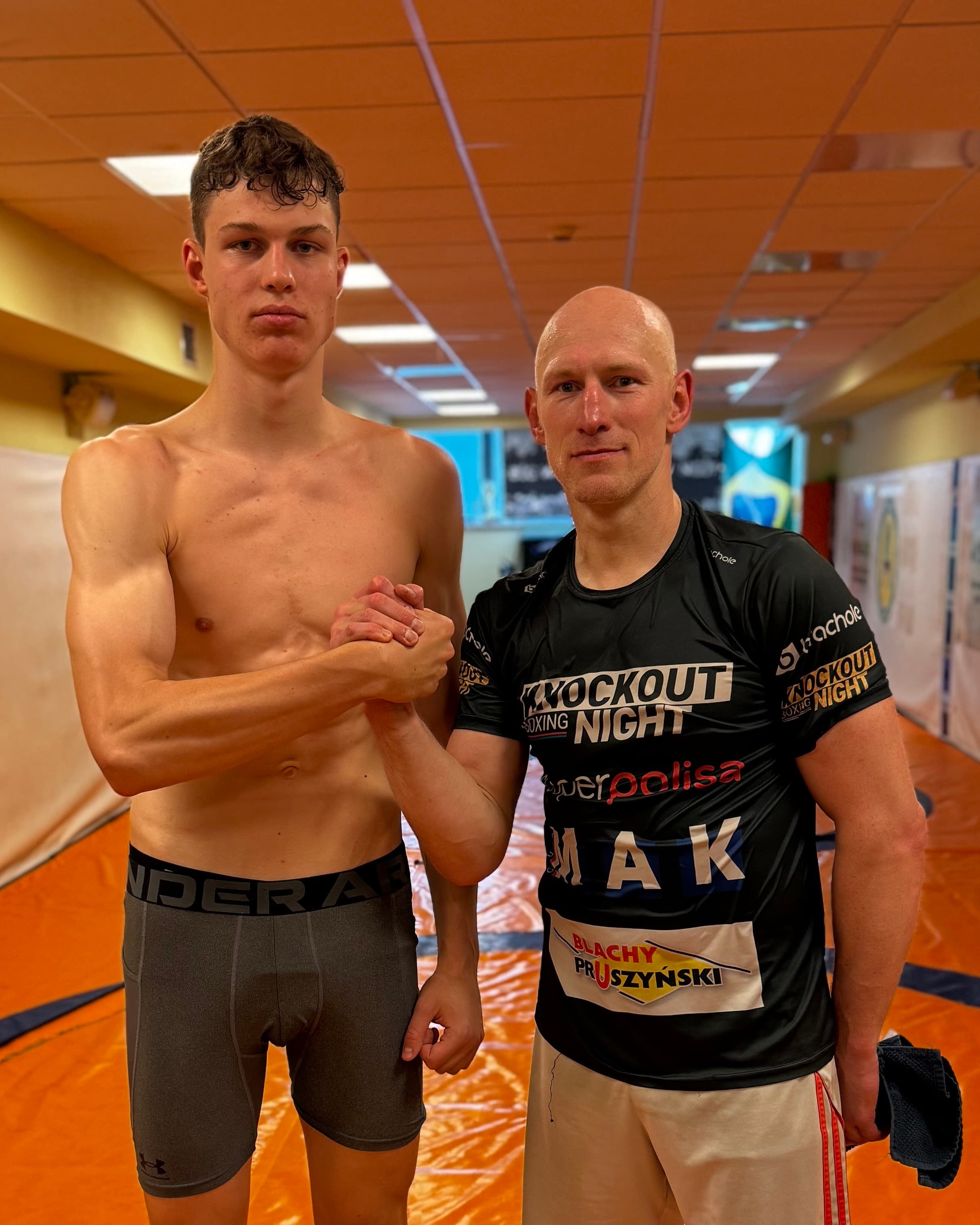 Aleksander Okniński ściska dłoń zawodnika w koszulce Knockout Boxing Night na macie zapaśniczej
