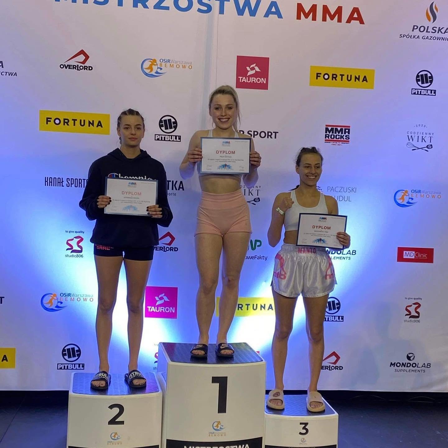 Zawodniczka na pierwszym stopniu podium z dyplomem, na tle banera mistrzostw MMA