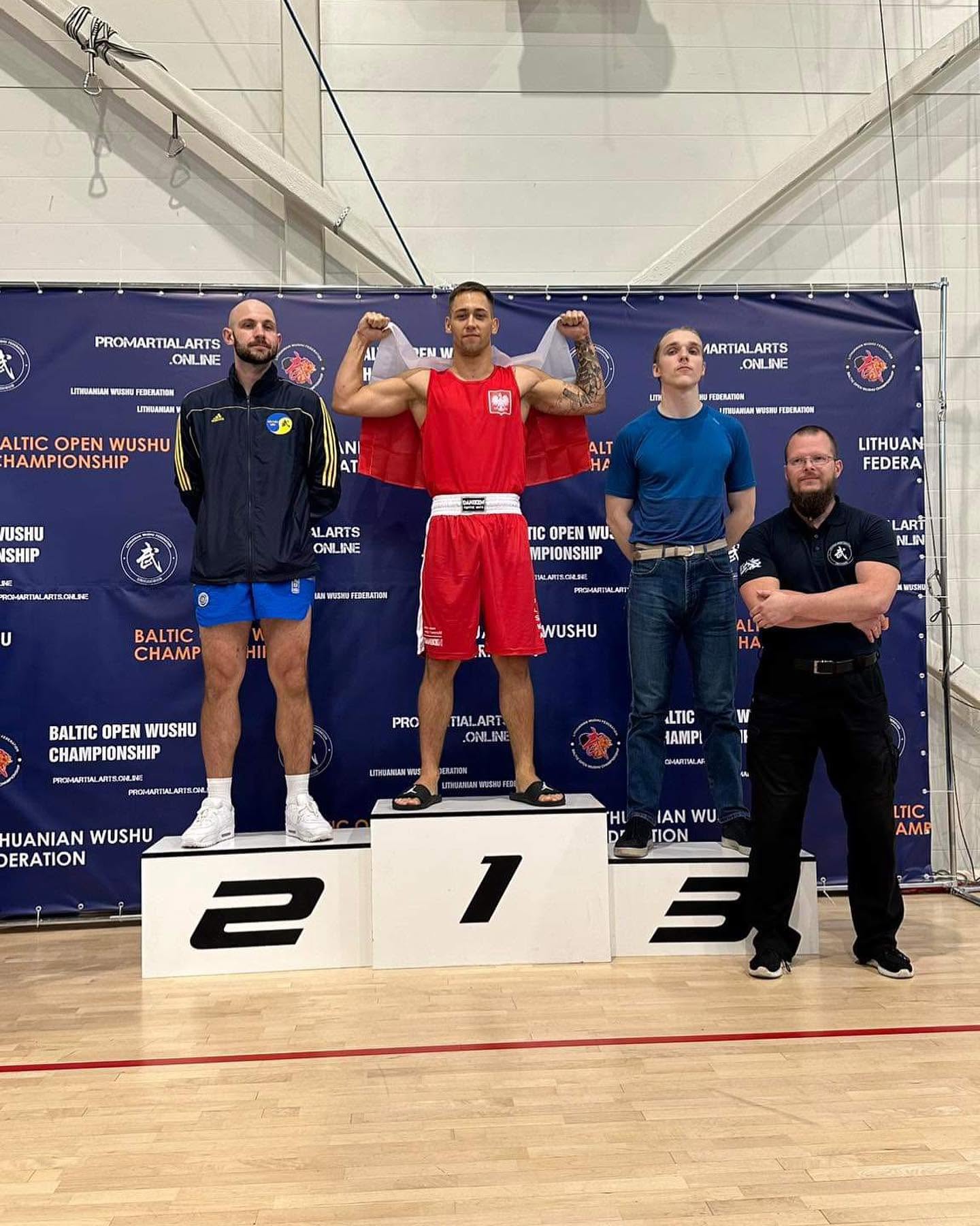 Zawodnik w czerwonym stroju na pierwszym stopniu podium, z polską flagą na ramionach, na mistrzostwach wushu